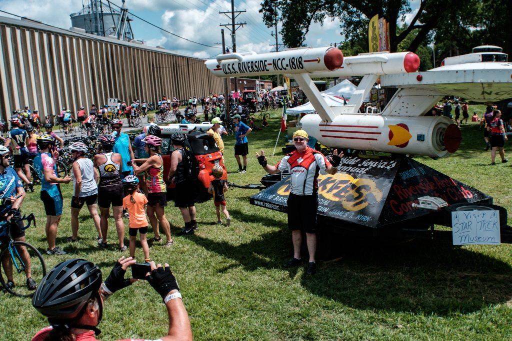 A RAGBRAI rider is seen posing with the miniature model U.S.S. Riverside in Riverside, Iowa, on the sixth day of the event, Frida7, July 27th, 2018. Riders rode from Sigourney to Iowa City on Day 6 of this years ride. (Jared Krauss/The Daily Iowan)