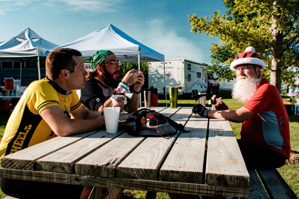 Ben Schwartz (L) of Appleton, Wisconsin, Tim Francis (L) of Davenport, Iowa, and Ed Feeney (R) of Dubuque, Iowa enjoying one of Mr. Pork Chops vittles on the Flynn family farm, just outside Wellman, Iowa during the sixth morning of RAGBRAI on Friday, July 27th, 2018. When asked how the ride was going, Ed said, The rain a couple nights ago just tore up my tent. I had to tie my poncho over top. Its kept together by clothespins and ducttape now. But it only has to last one more night. Riders rode from Sigourney to Iowa City on Day 6 of this years ride. (Jared Krauss/The Daily Iowan)