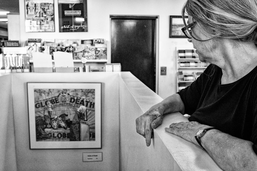 Nana Burford, a painter and employee at Prairie Lights Books, looks on to a painting, Globe of Death, completed by her father, Byron Burford, in 1967 inside Prairie Lights Books in Iowa City, Iowa on Wednesday, August 22, 2018. The painting was originally purchased by Jim Harris, the founder of Prairie Lights Books. She remembers him coming over to her fathers house and going through his studio. She said, My father loved the circus. He traveled with them and played and the drums, and he painted the side show displays. Nana said she swore she would never become a painter, but now shes moving apartments so she can have a studio. Her mother, too, was a painter. But her style is not overly impacted by either she said, My parents bent over backwards to make sure they didnt influence me.