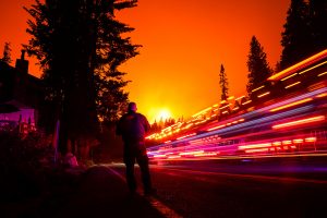 A vehicle streaks by in a long exposure as Fresno County Sheriff Deputy Jeffery Shipman stands along CA-168 as the Creek Fire creeps closer to town on Sunday, Sept. 6, 2020 in Shaver Lake, California. (Kent Nishimura/Los Angeles Times/TNS)