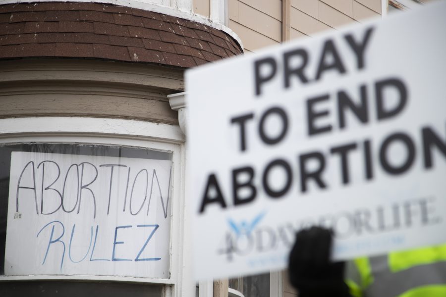 A sign reading “ABORTION RULEZ” is seen in a window behind a pro-life protester during an abortion protest and counter protest outside of the Emma Goldman Clinic in Iowa City on Saturday, Jan. 21, 2023. (Matt Sindt/The Daily Iowan)
