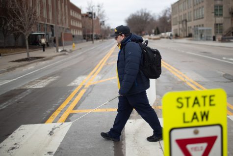 Jerad Nylin crosses the street with his white cane by the Main Library on Feb. 23, 2023. Nylin is a blind student attending the University of Iowa. Nylin wears a navy blue coat with a yellow jacket underneath and a black Hawkeye baseball cap.