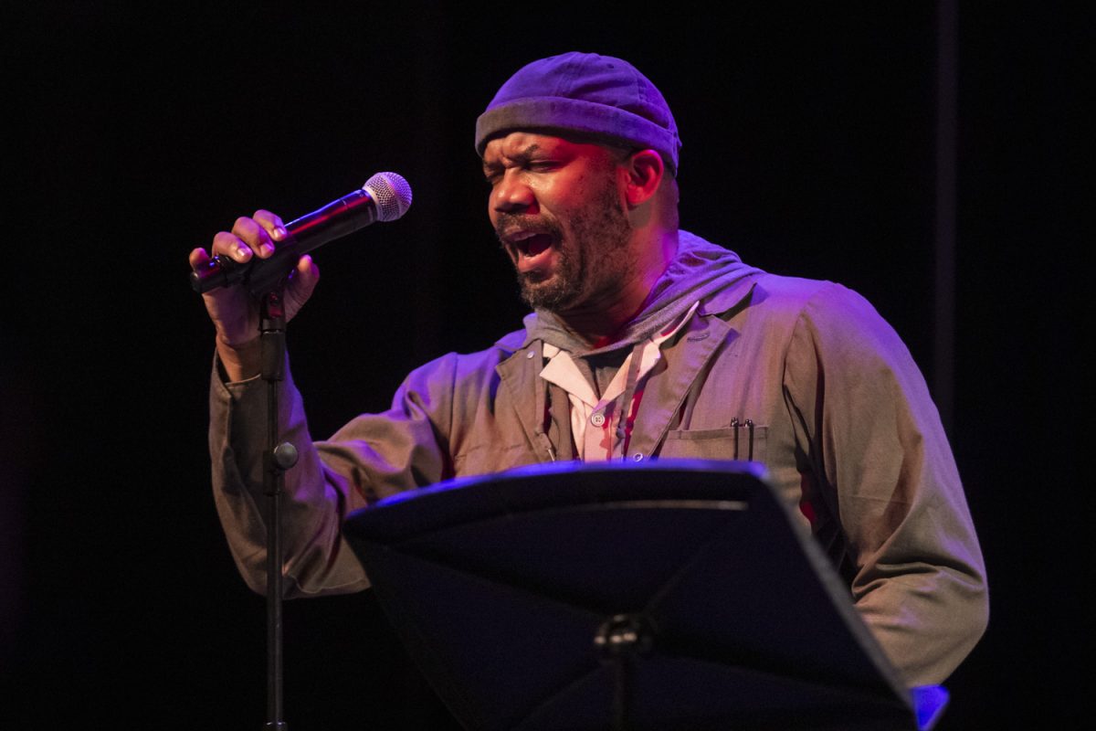 Emanuelee “Outspoken” Bean performs an original composition at Mic Check Poetry Fest at Hancher in Strauss Hall on Friday, Nov. 8, 2024. “I wear coveralls or overalls when I do a performance because it is a living metaphor for the ones who work,” he said during his act. Along with Bean, the event featured spoken word poetry from Rudy Francisco and teens from IC Speaks.