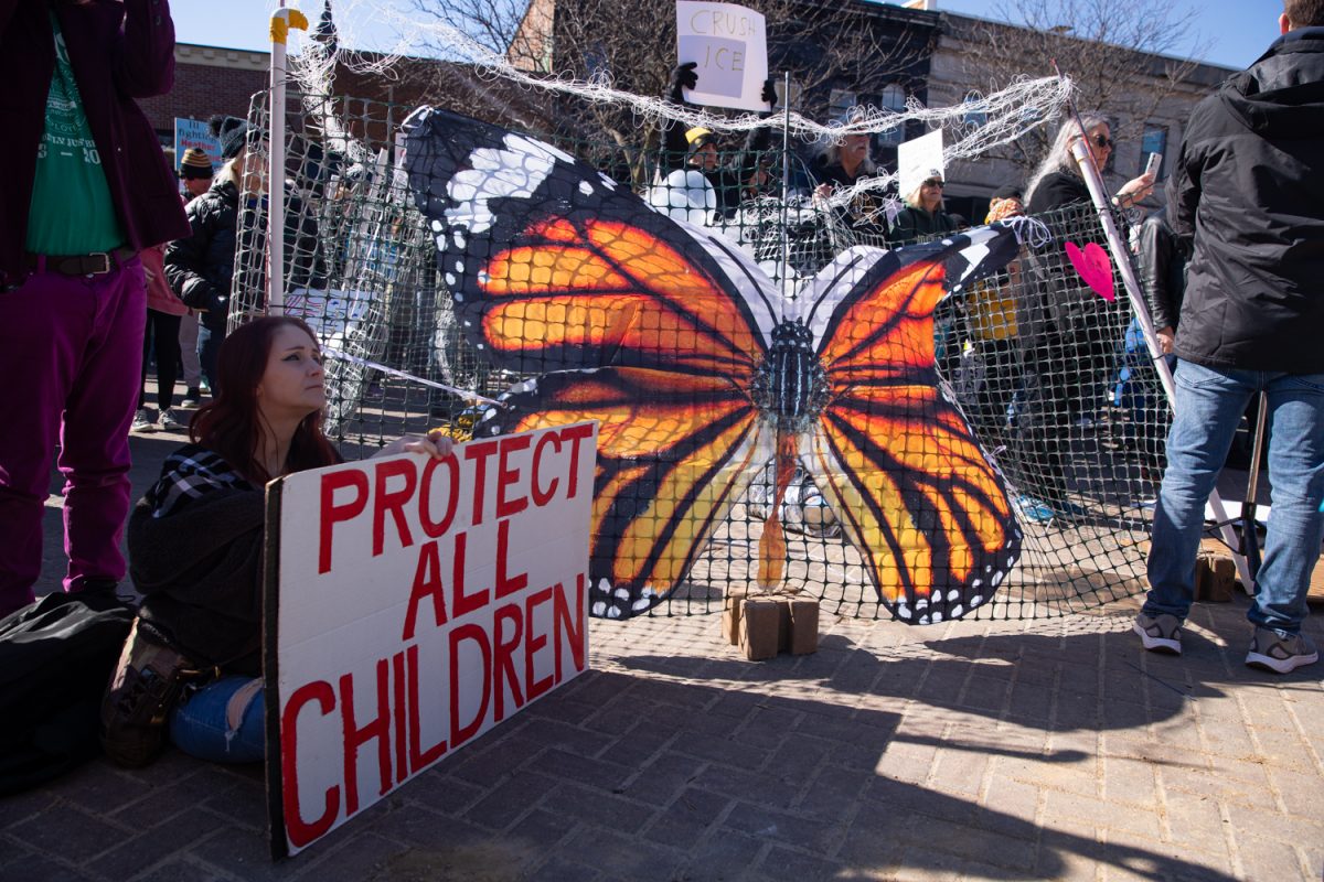 A protester sits with a sign beside an art installation symbolizing ICE detention camps during a “No Kings” protest in downtown Iowa City on Saturday, March 28, 2026. The Iowa City demonstration is part of protests taking place nationwide against President Donald Trump’s administration, ICE, and the war in Iran.